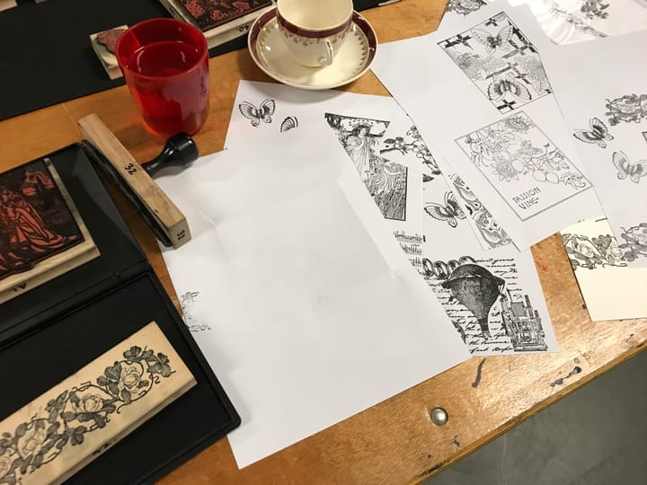 Table spread with art stamps, ink pad, paper sheets with stamped images, a red glass and a teacup.